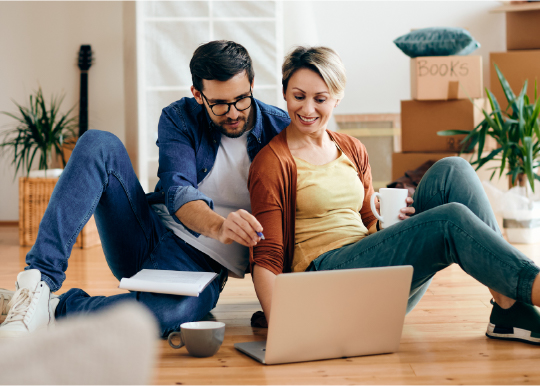 Couple sitting on floor of home looking at laptop. A window with stacked cardboard boxes is in the background.